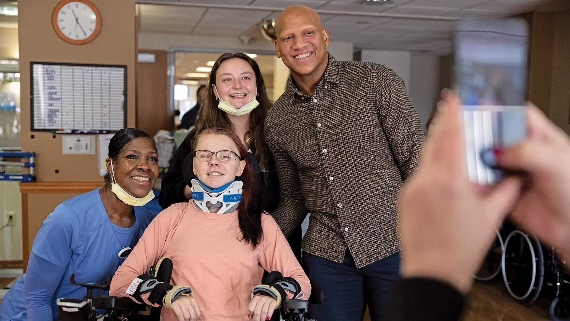 A young woman in a neck brace leans back in her wheelchair and looks at a person taking a cellphone photo, who’s barely shown at the edge of the photo. On the patient’s arms are braces, and her fingernails are painted. Around her are Ryan Shazier and two hospital workers also posing for the camera.