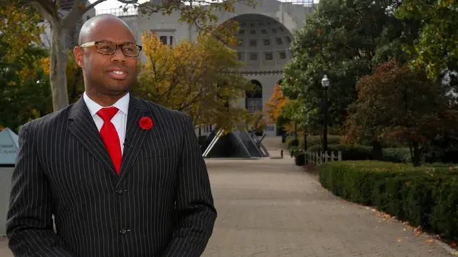 Dressed in a pinstripe suit and wearing glasses, a Black man stands in front of Ohio Stadium, looking into the distance and smiling