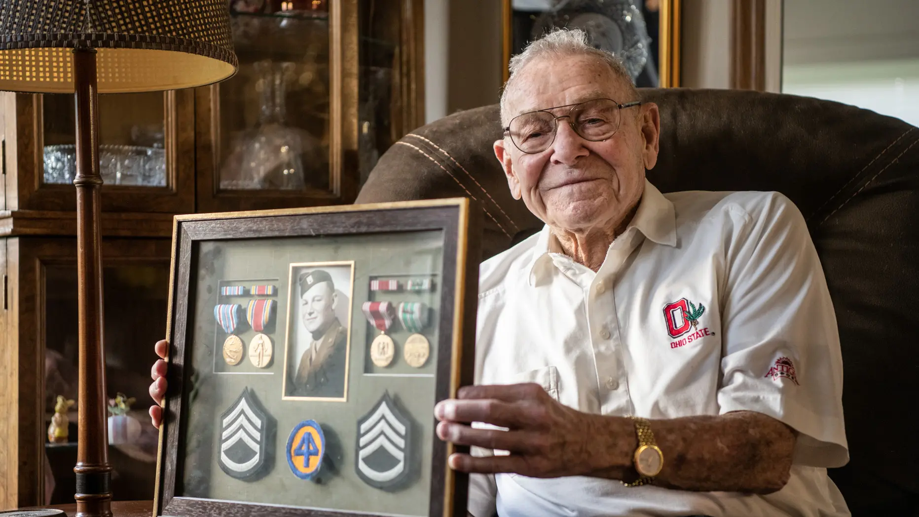 Merle Lashey Jr. shows a framed collection of military patches with his Army photo, taken when he joined to fight in World War II. He wears an Ohio State-emblem shirt and sits in a leather chair with a small, proud smile.