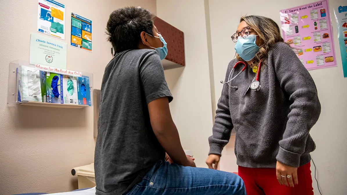 A young doctor in a surgical mask prompts a child patient to raise his shoulders during a medical exam