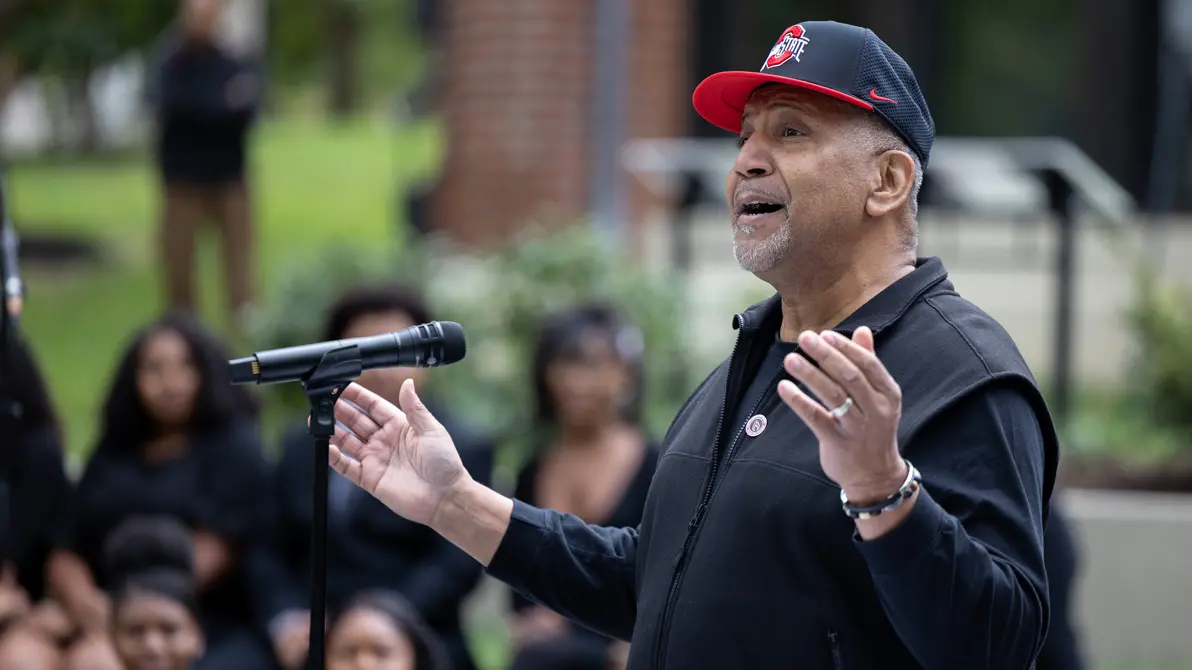 A Black man wearing an Ohio State ball cap gestures with hands spread wide as he speaks to an audience--only vaguely suggested in the background--at the dedication ceremony. He looks optimistic and proud. 