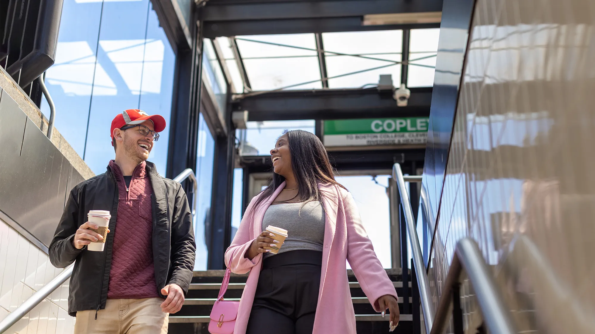 Josh Javor, a white man with glasses and a baseball cap, and Dominque McClean, a Black woman with long hair in a dress coat, smile at each other as they walk down stairs into a subway entrance.