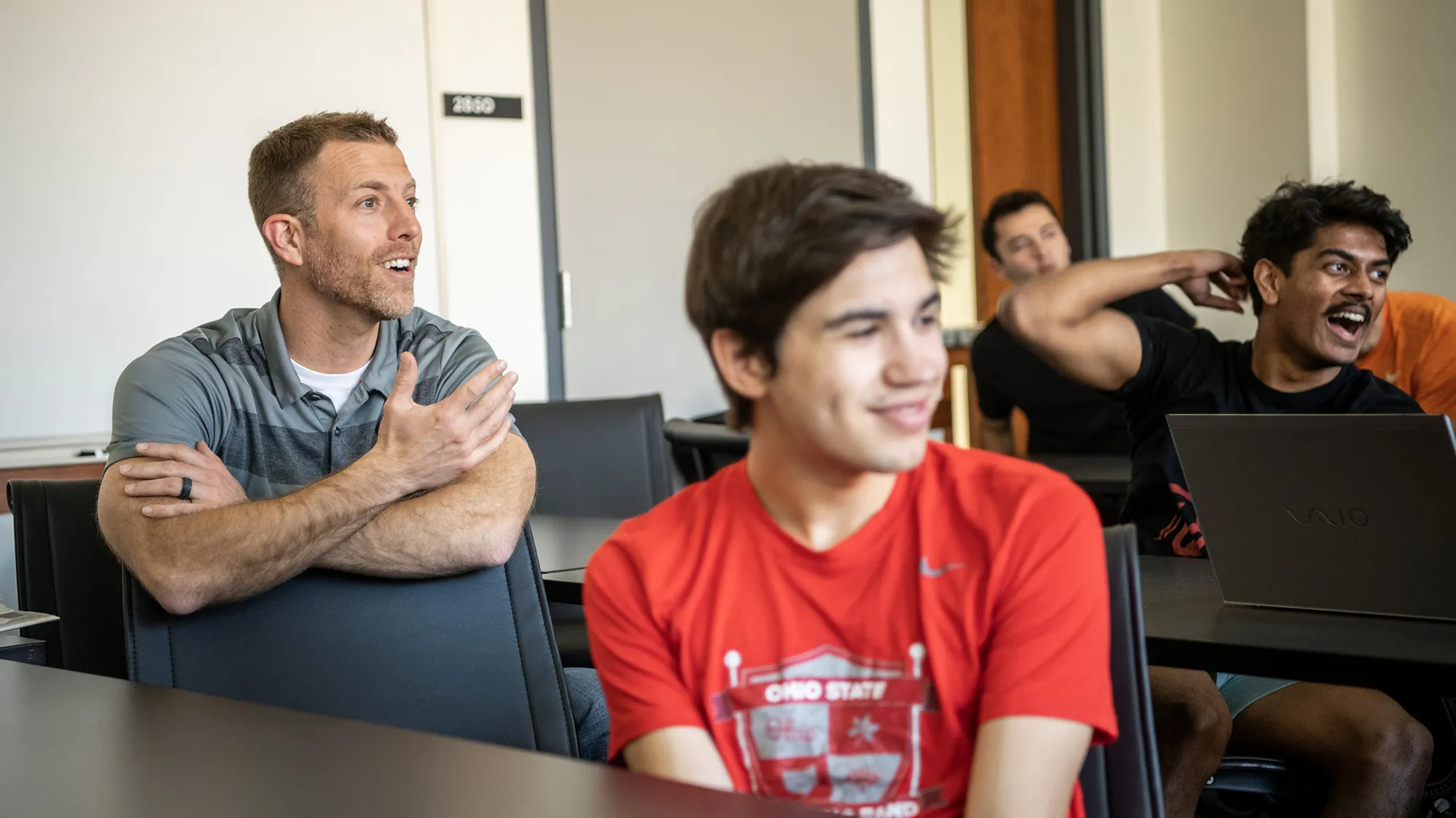 In a classroom, a group of men are having a good time. Teacher Chad Zipfel, a white man with short hair and a beard scruff, leans on the back of a chair in front of where he’s sitting. He’s sitting among students and talking, gesturing and smiling as his eyes shine and he looks at someone out of the photo frame. The students around him, looking in the same direction, also laugh or smile.