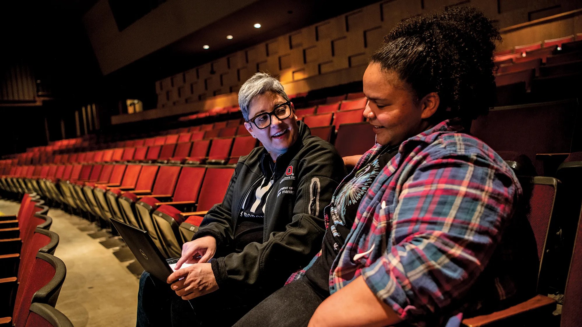 Sitting at the end of a row of chairs in a theatre, teacher Sherée Greco, a white woman with short gray hair and thick-rimmed glasses, points to a laptop screen she’s holding and talks with a student, a young Black woman with a curly pony tail. They’re angled slightly toward each other and both are smiling — the younger woman is looking down as if she’s listening intently and perhaps slightly embarrassed but pleased by what’s being said.