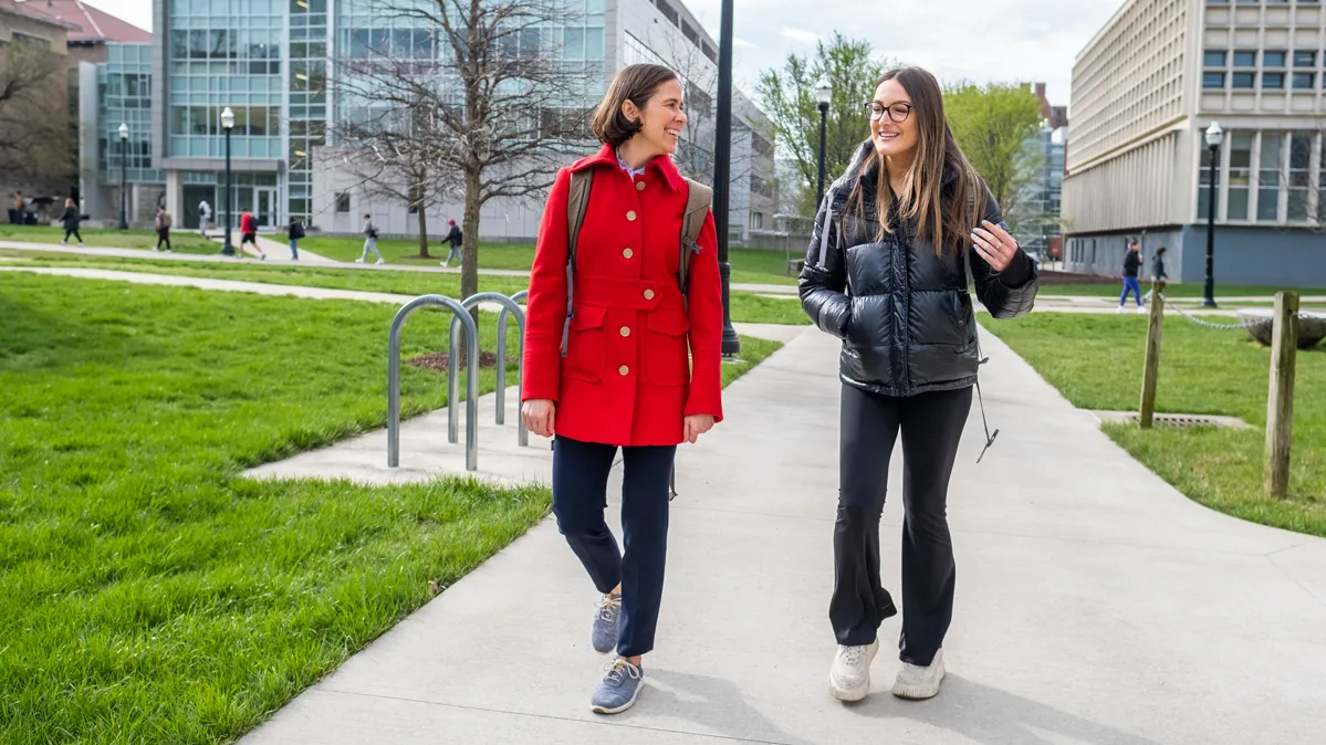 Carmen Swain, a white woman with short hair, and Joie Platt, a young white woman with glasses and long hair, smile and chat as they walk down a sidewalk on campus, both wearing sneakers. Though they wear winter coats, the grass is green and the light is bright. Behind them are two classroom buildings whose fronts are mostly windows.
