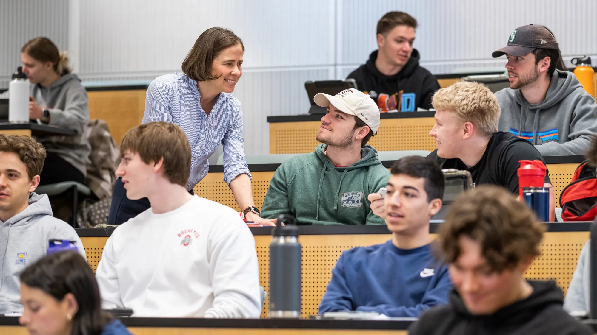 In a classroom with mostly male students, teacher Carmen Swain leans on a desk to talk to two students. She and the young men seem relaxed and are smiling.