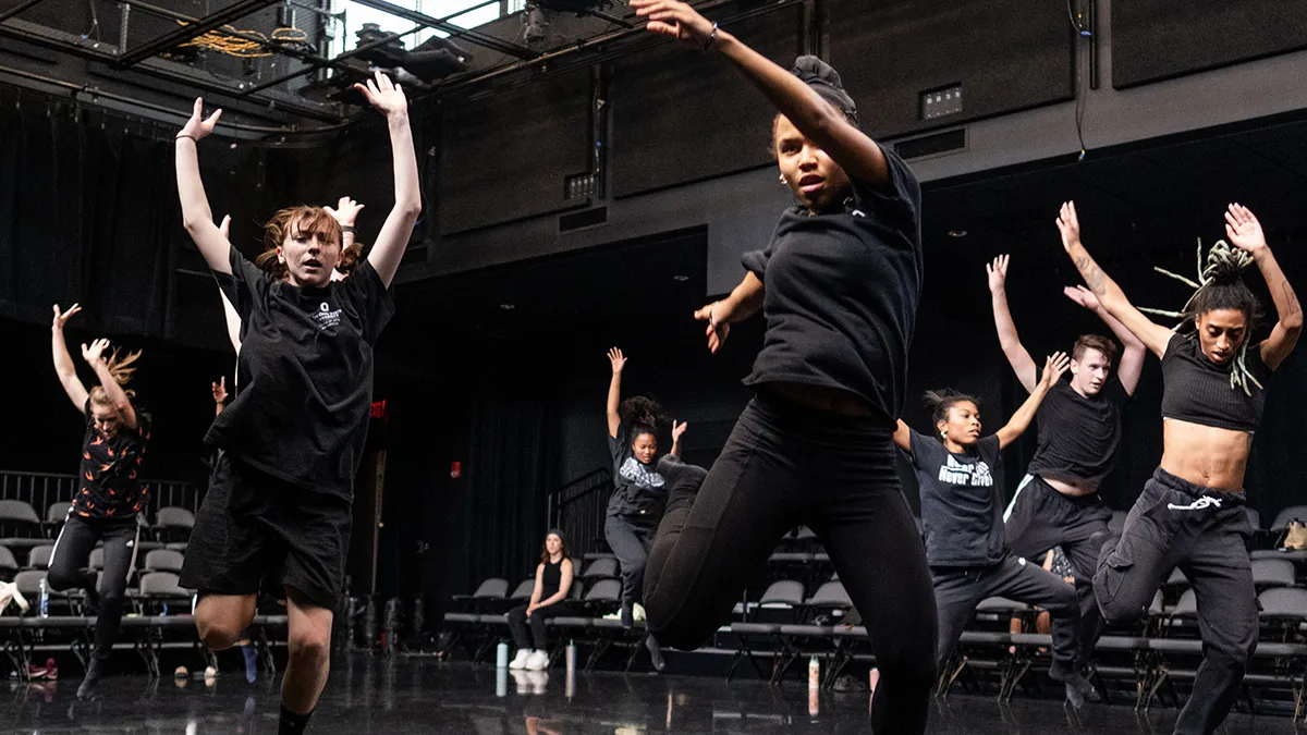 Seven dancers wearing black clothes—the color being the clothes’ only similarity—leap into the air with arms extended above their heads as they enact a scene from the island in “The Tempest.” 