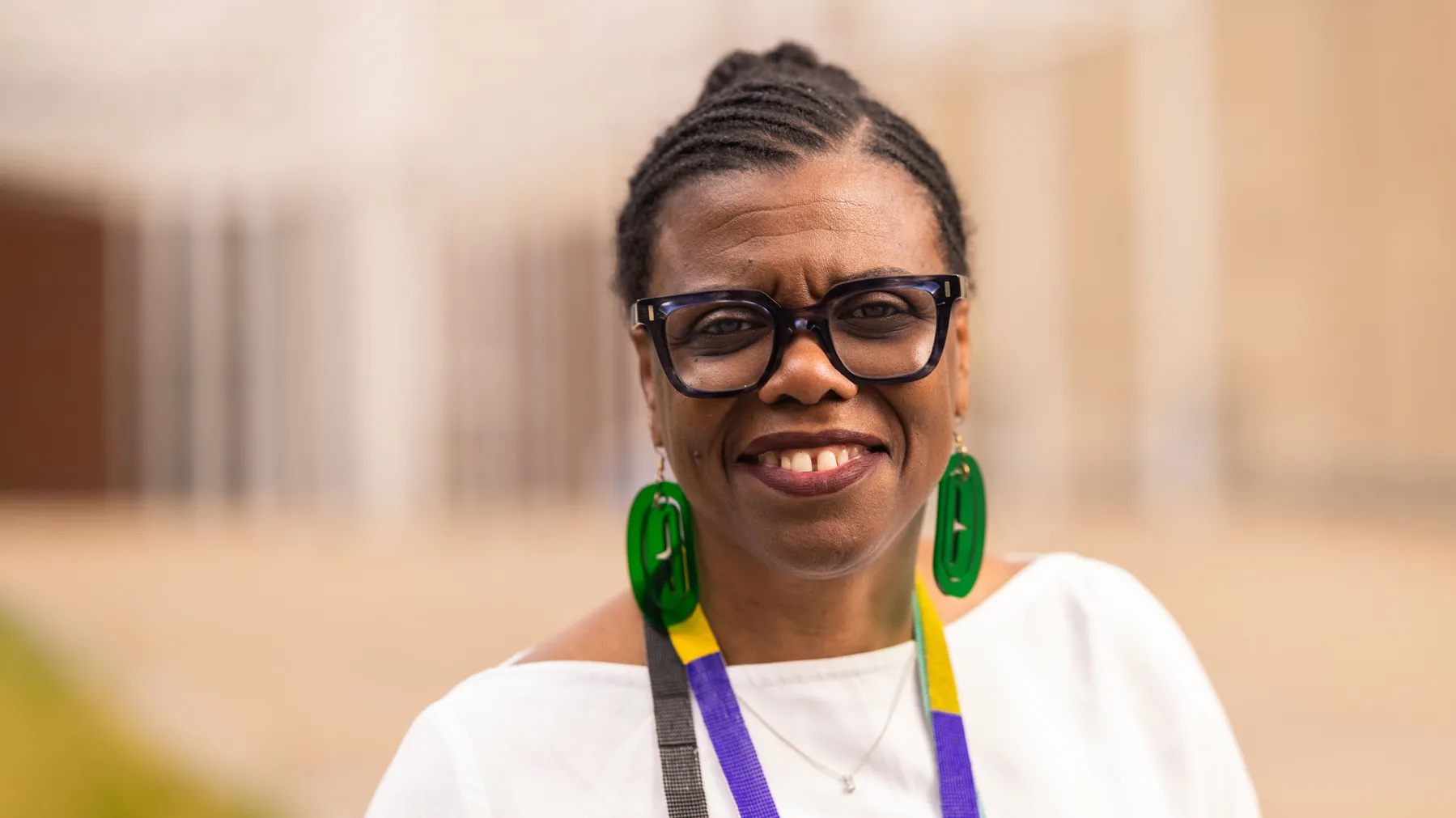 Gaëtane Verna, a Black woman with her braided hair pulled back in a bun, is shown from the shoulders up, smiling as she seems to make eye contact with the viewer outside of the Wexner Center for the Arts. She wears thick-rimmed glasses and big transparent earrings, adding to her confident, artsy presence.