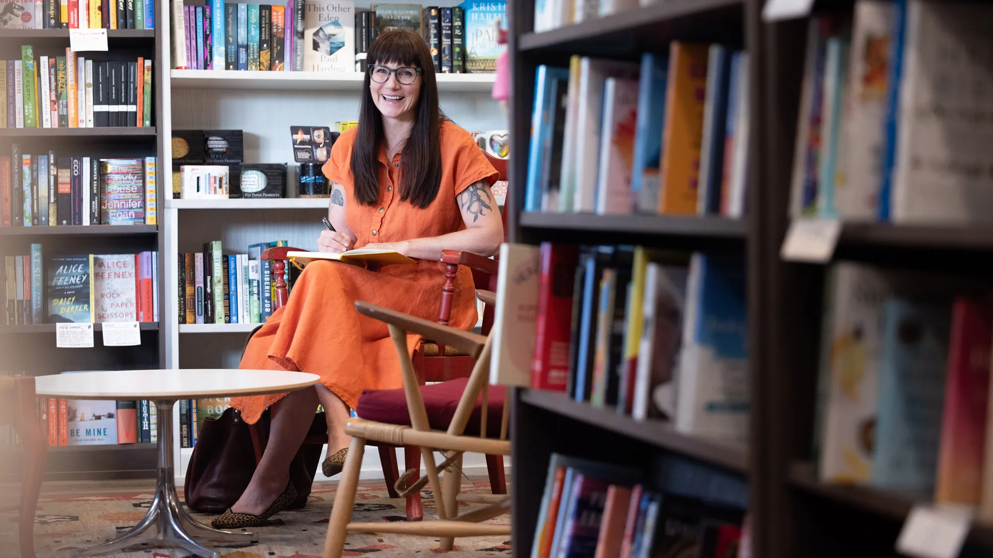 Maggie Smith — a white woman with long brown hair, glasses and tattoos of lemons and leaves on her upper arms, which you can see because she’s wearing a linen, short-sleeved dress —smiles as if she’s laughing or talking as she sits in a painted wooden chair. On her lap is a book she’s writing in and behind her are more books on shelves.