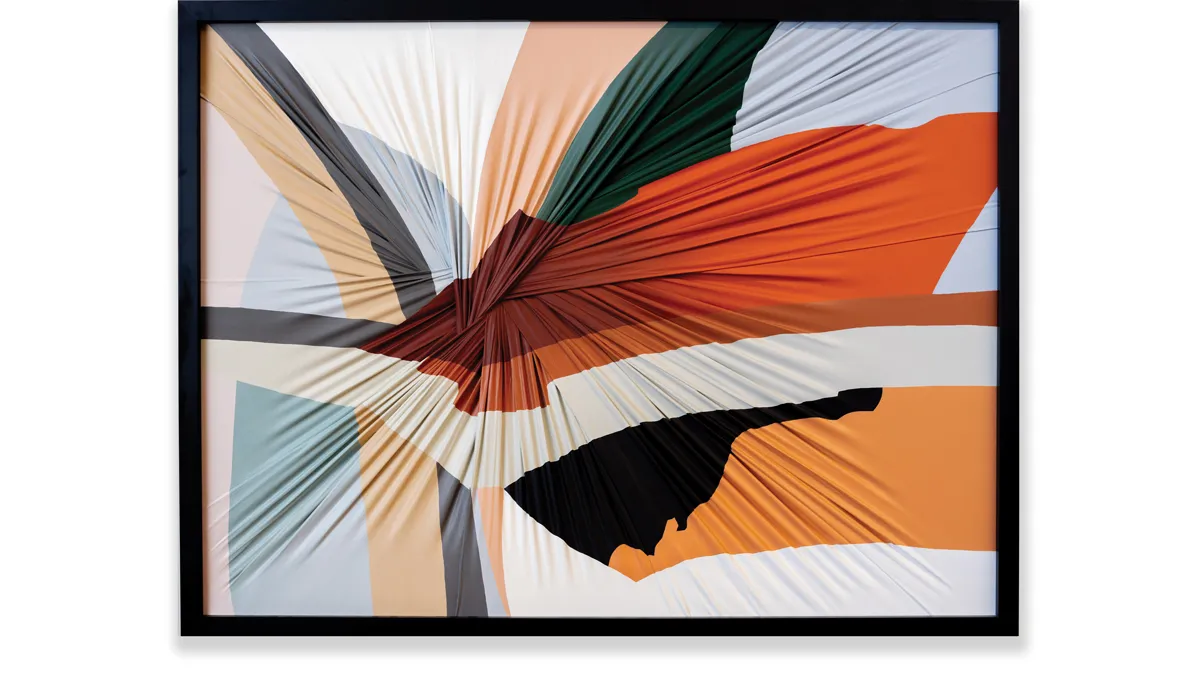 A framed piece of fabric, which looks as if the center was tied into a knot and then the whole piece flipped, represents the colors of costumes from a 2021 play. They are both warm and cool colors; orange shades and light blue stand out.