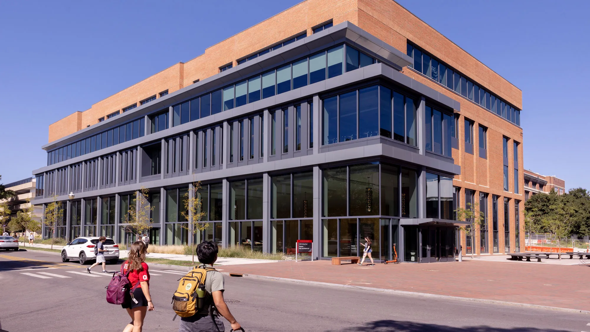 A building that is much longer than it is tall has a facade with two levels of windows and warm colored brick. It looks modern and ready for great things to come from within. Two students cross the street toward the building.