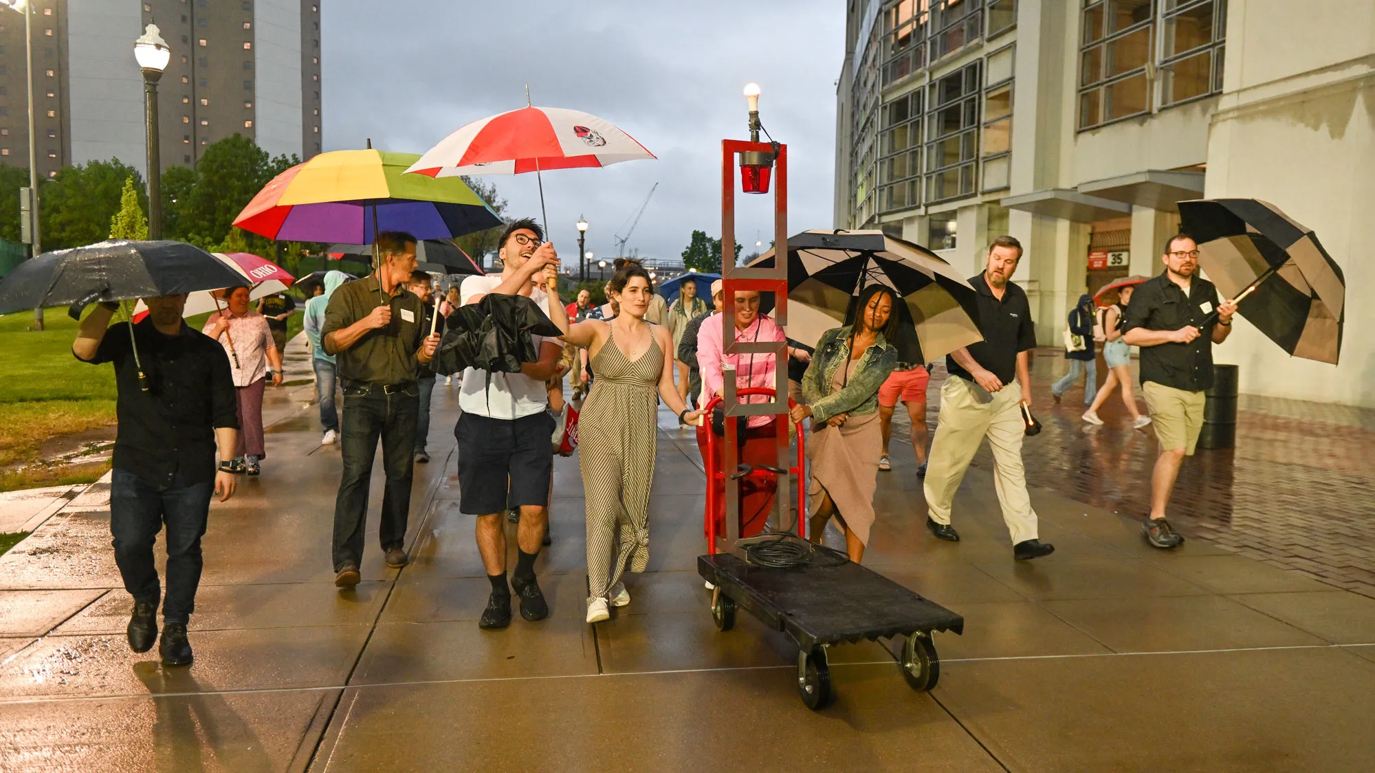 A crowd of teachers and students, many carrying umbrellas, accompany a rolling cart with a large metal OSU sculpture (those letters are stacked on top of each other) with a light bulb at top — a ghost light. As it rains, the people pass by university buildings in an informal parade.