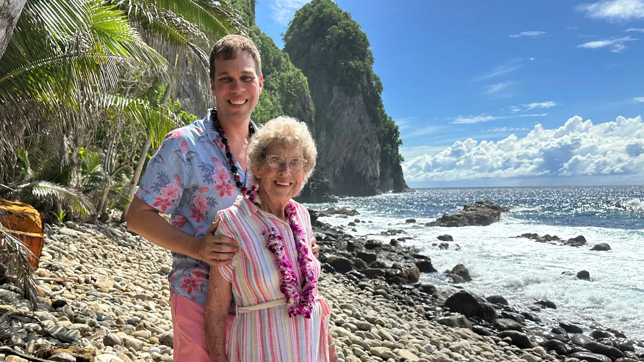On a beach of palm-sized rocks (not sand), a man stands behind his grandma with his hands on her shoulders. They both wear leighs — hers made of flowers; his, nuts — and dress in tropical clothes. They seem happy there between the ocean and palm trees.