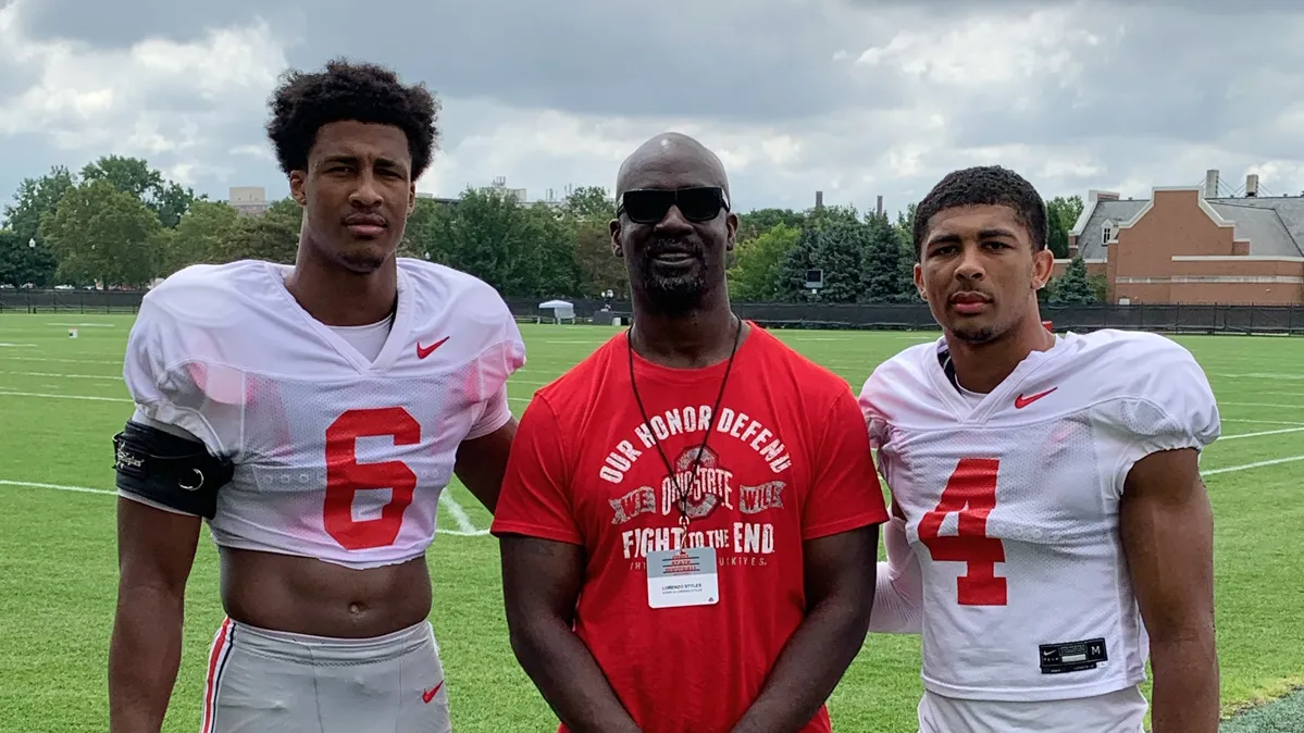 Standing in order of tallest to shortest on the football team’s practice field, Sonny, Lorenzo C. and Lorenzo P. Styles pose for a photo. The young men are on the ends, with serious expressions as they look at the camera, and their dad is smiling proudly between them. Both sons have a hand on his back and hold their helmets in their outside hands. Their dad has his hands clasped in front of him. 