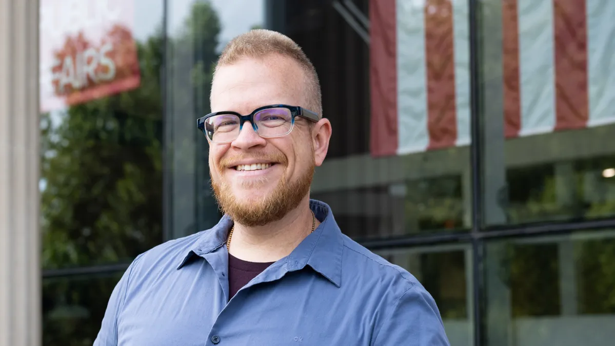 David Hibler, a broad-shouldered white man with close-cropped hair and beard and thick-frame glasses, leans on a railing outside Ohio State’s John Glenn College of Public Affairs. He has a friendly smile, the kind that crinkles his eyes, and in the background, light shines off the building’s windows. An American flag is visible depending on the reflection.