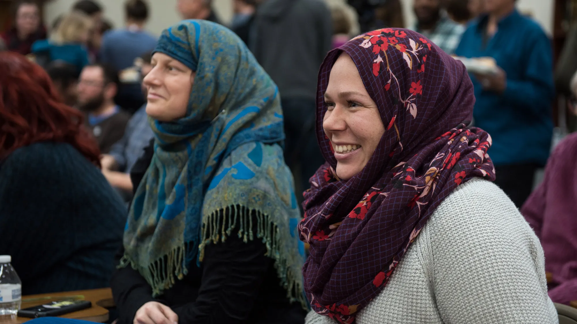 Gretchen Klingler wear a purple headscarf and smiles as she listens to someone out of the photo. Beside her is a white woman wearing a blue paisley headscarf and people surround them chatting with one another. 