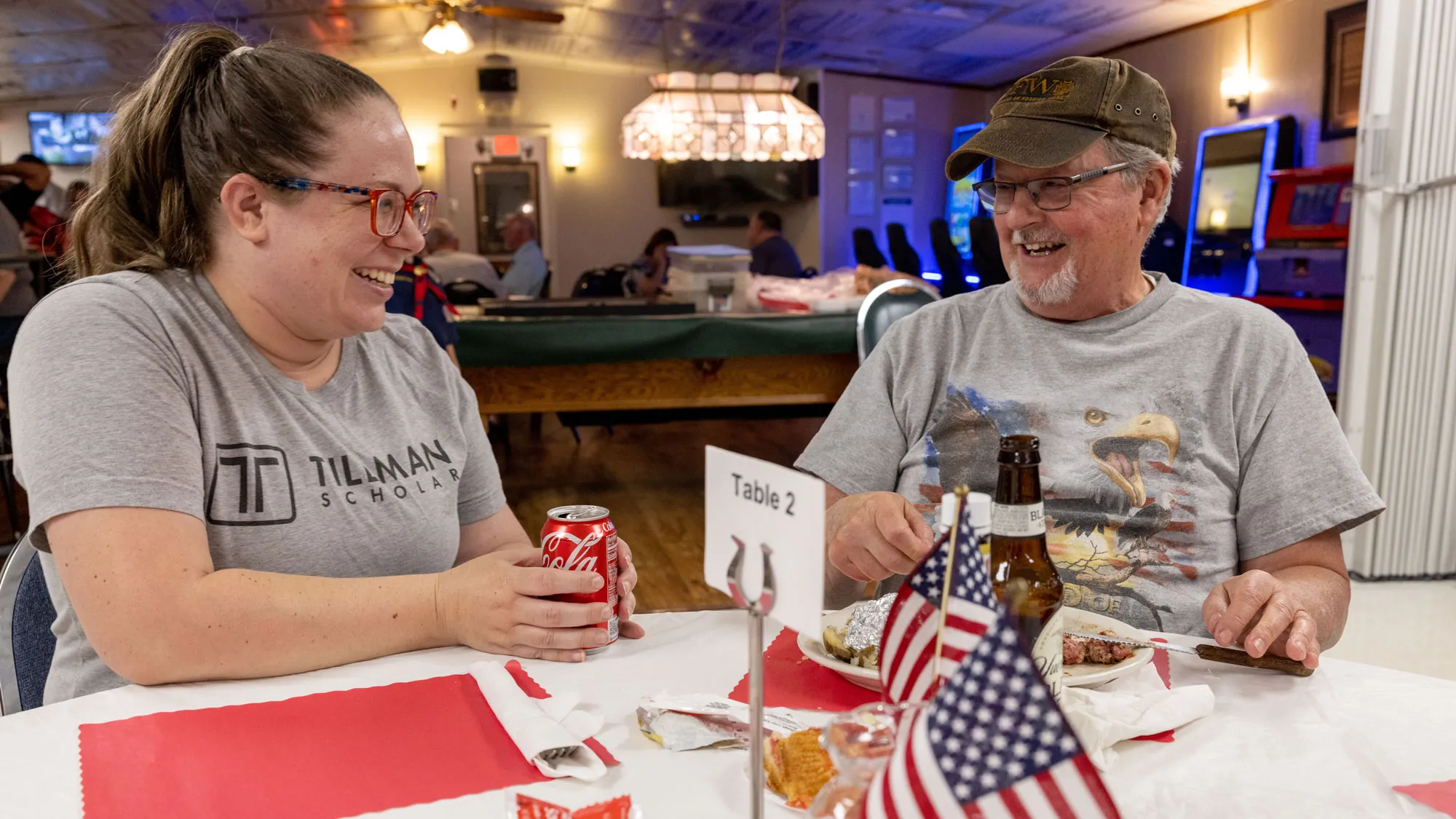 Wearing a T-shirt that says Tillman Scholar and laughing, Gretchen Klingler shares a table and chats with veteran George Walker, a gray-haired white man, at her VFW post. He’s laughing, too, and his T-shirt has a bald eagle. In front of him is a plate of food and American flags make the table centerpiece. Behind them, more people sit at tables. 