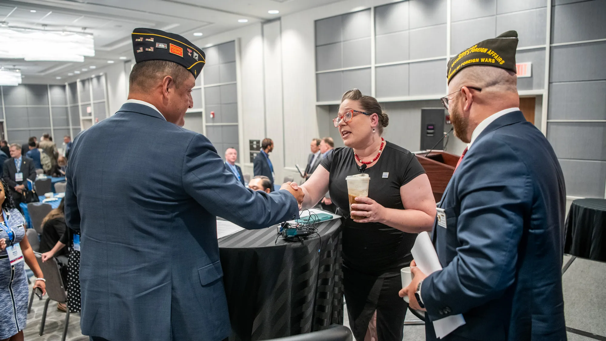 At a table on a small stage, Gretchen Klingler, a white woman with a style reminiscent of the 1940s, leans across a table to shake hands with a white man wearing an American Legion uniform cap and a suit. They’re making eye contact, and next to them smiling is a white man in a suit, this one with a VFW uniform cap. 