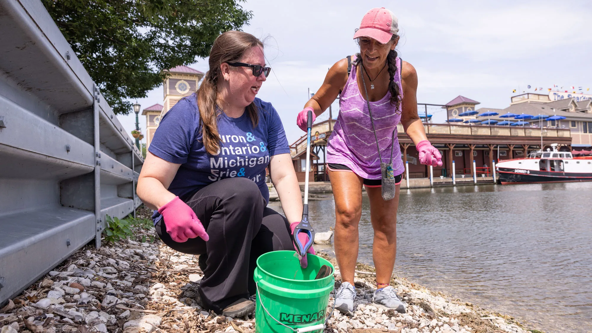 Between Lake Erie and a short metal wall, two women pick up trash on a rocky shoreline. They have a bucket to put trash in and wear gloves. One woman crouches down the other leans toward her with a pickup tool claw on a long handle. 