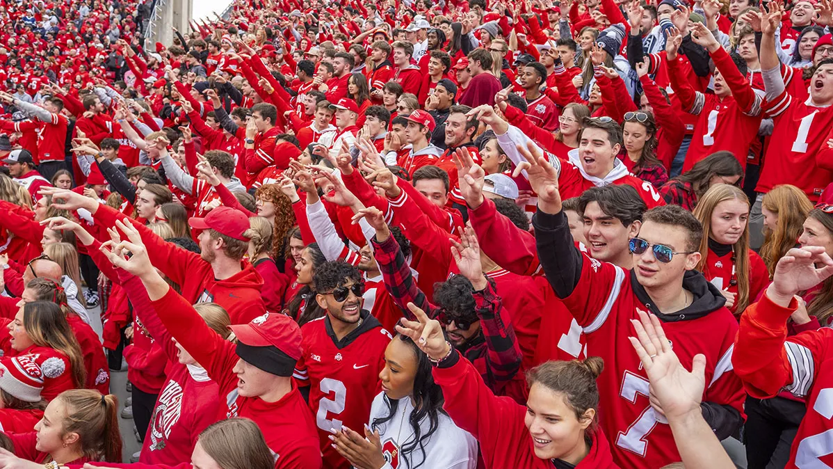 A sea of students in the stands at a football game wear Ohio State jerseys as they laugh and reach out while cheering on their team.