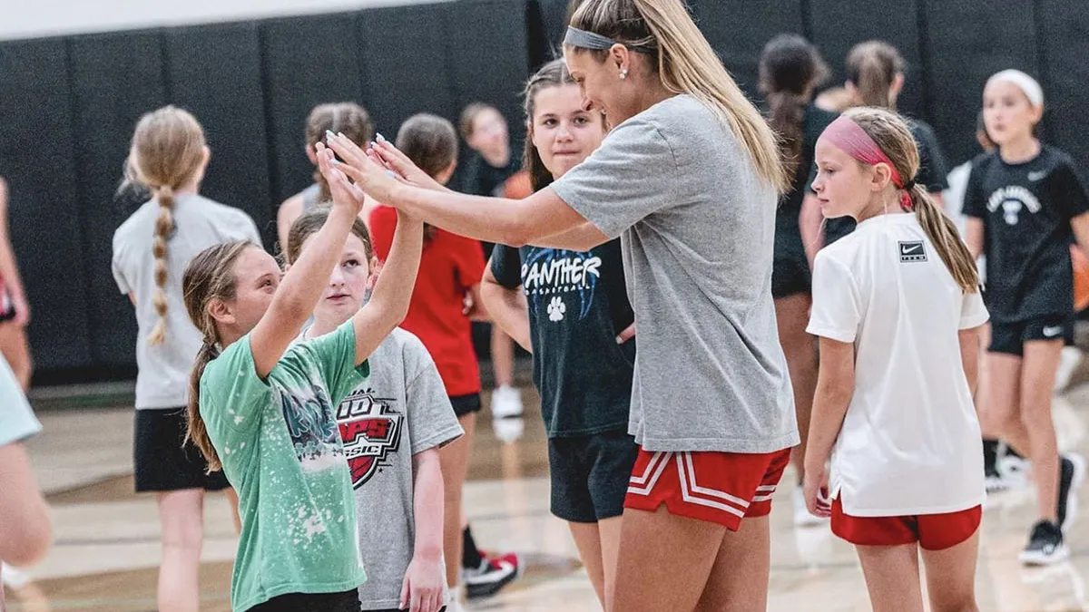 A tall young woman with a long blond ponytail gives a child a high five as other young players mill around her in a gym. They’re all wearing workout clothes and are in the midst of practicing basketball.
