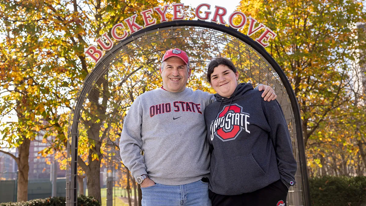 A smiling dad wraps an arm around his daughter’s shoulders. They both wear Ohio State sweatshirts and pose before the Buckeye Grove gateway, which spells out that name on the arch above their heads. Sunshine filters through the trees in the background, whose leaves have changed colors for fall.