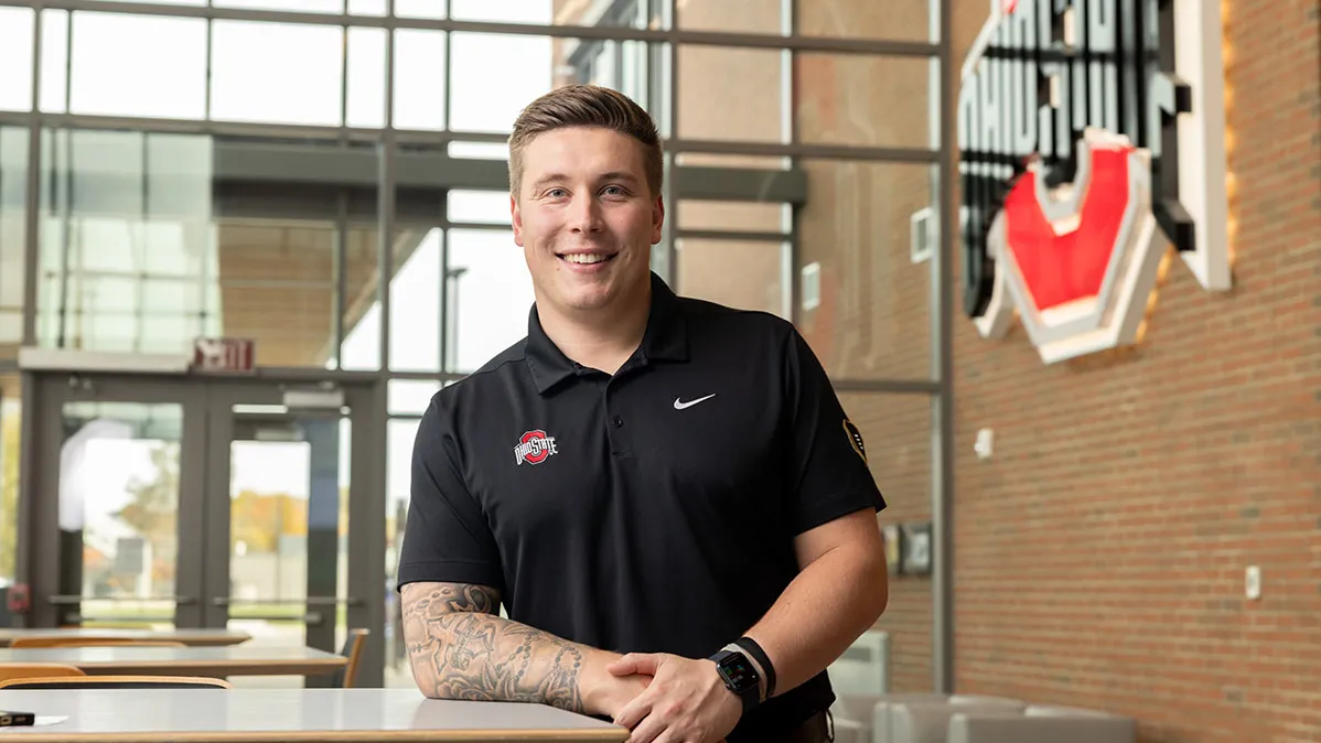 A young man wearing an Ohio State polo short smiles as he poses for this photo, leaning on a waist-height table in a building foyer where a large Ohio State logo hangs on the brick wall. He has neatly styled hair and tattoos on his right arm.