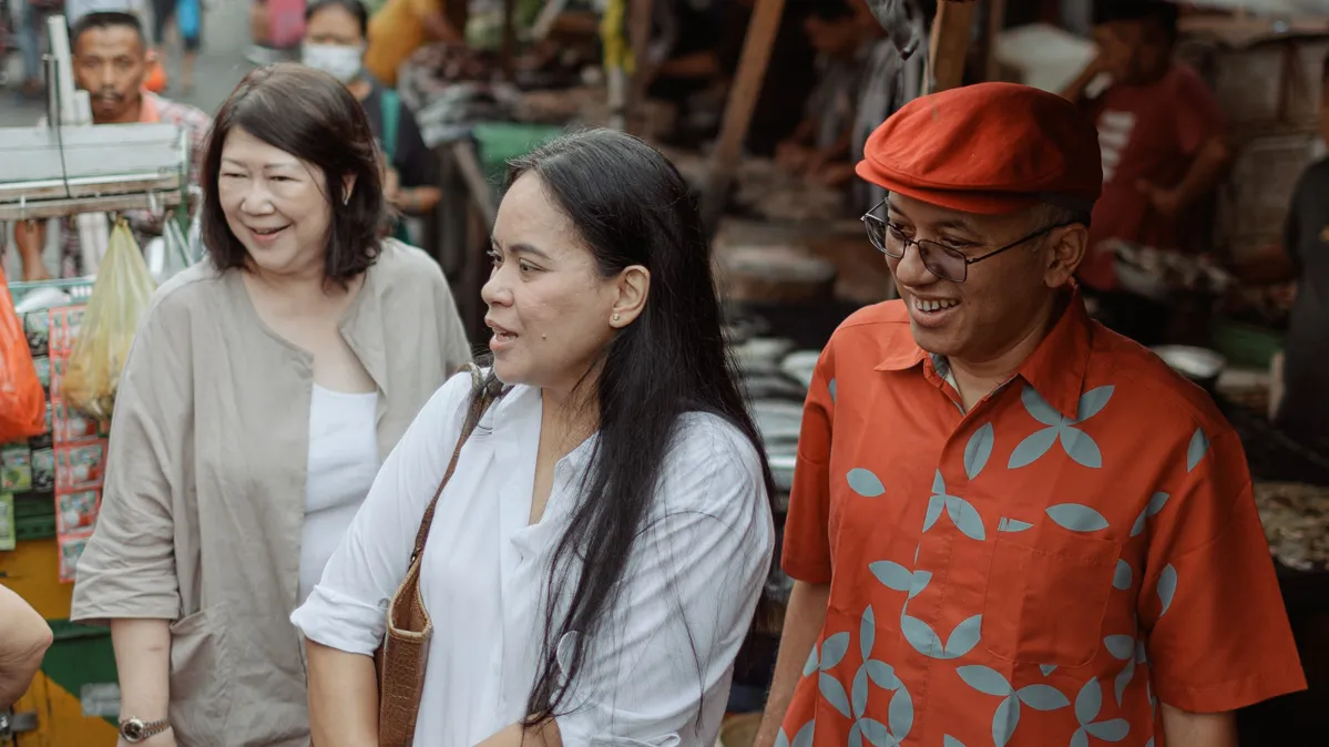 On a bustling street lined with stalls, three people take in an off-screen option. The man and two women smile as one of them comments on what it is they’re looking at. 