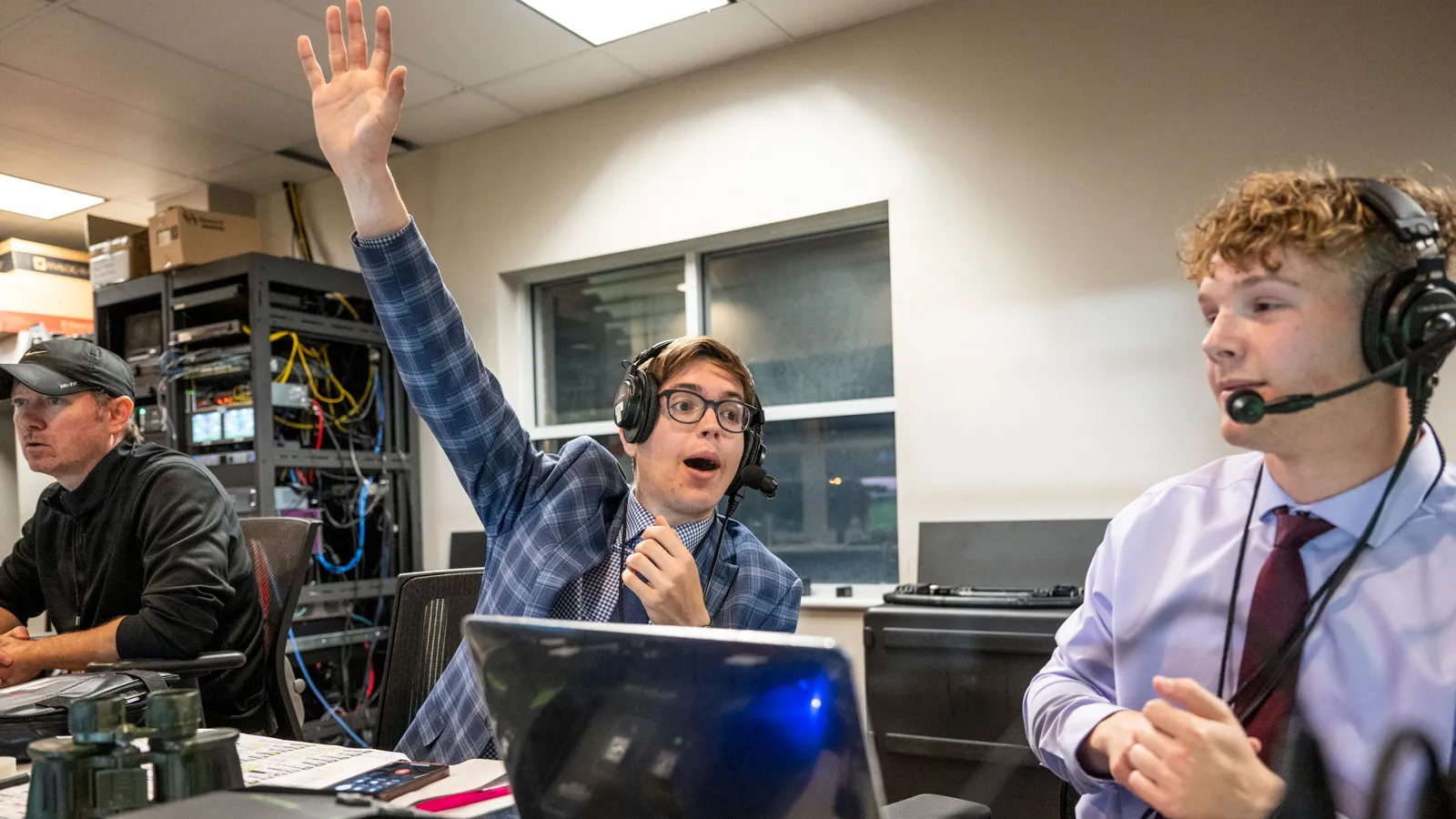 Dressed in a checked suit coat, Tyler Danbury reaches high as he sits behind a computer in the press box at a sporting event. On both sides are more journalists, and binoculars sit on the desk in front of him.