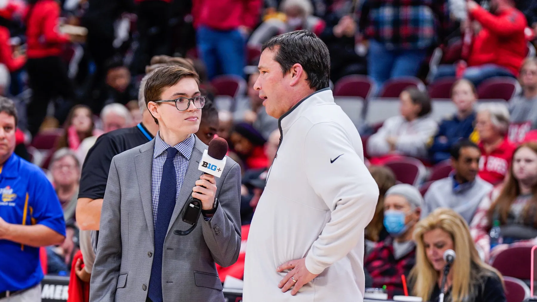 Standing on the sidelines of a basketball court, a young man in a suit and glasses listens intently and holds a microphone for the man he’s interviewing. That man, as adult in gym clothes, talk with hands on hips. Behind them, the stands are full with spectators wearing Ohio State colors.