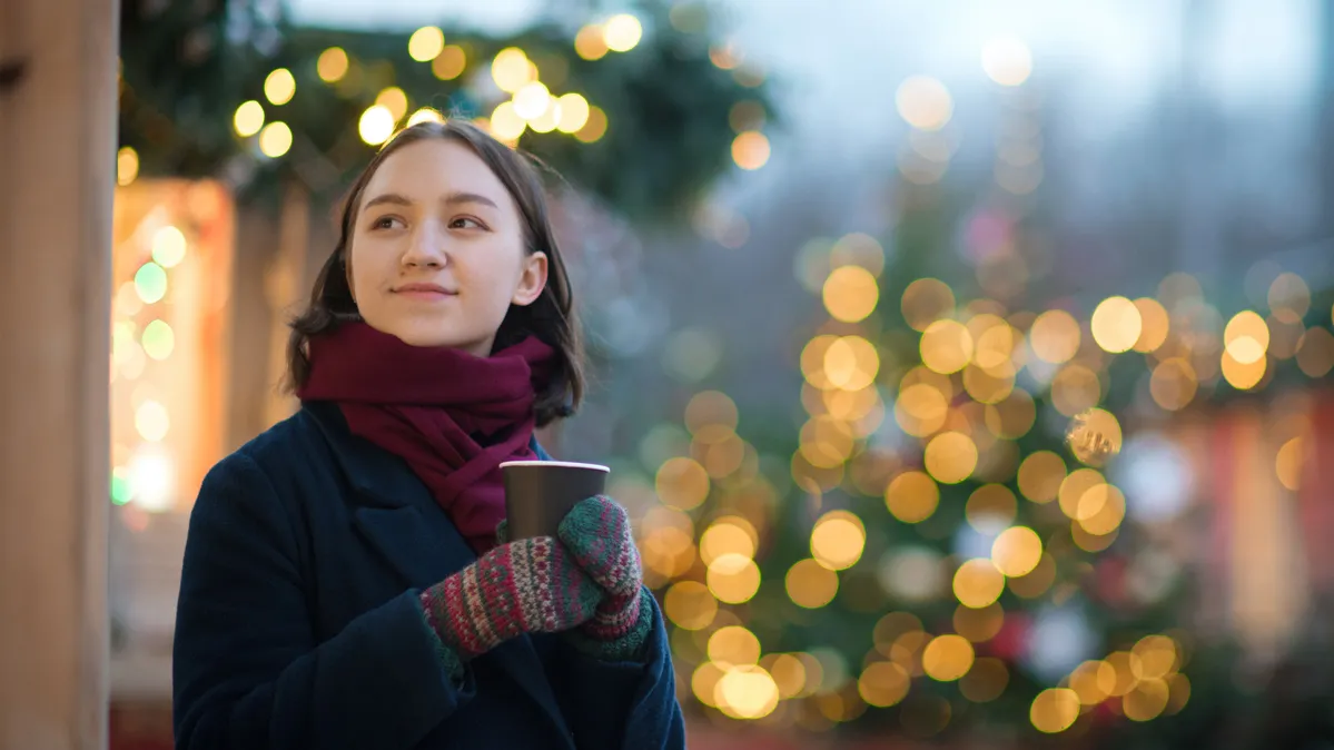 A young woman with a tightly bundled scarf and very slight smile stands among outdoor holiday lighting and holding a beverage cup with mittened hands