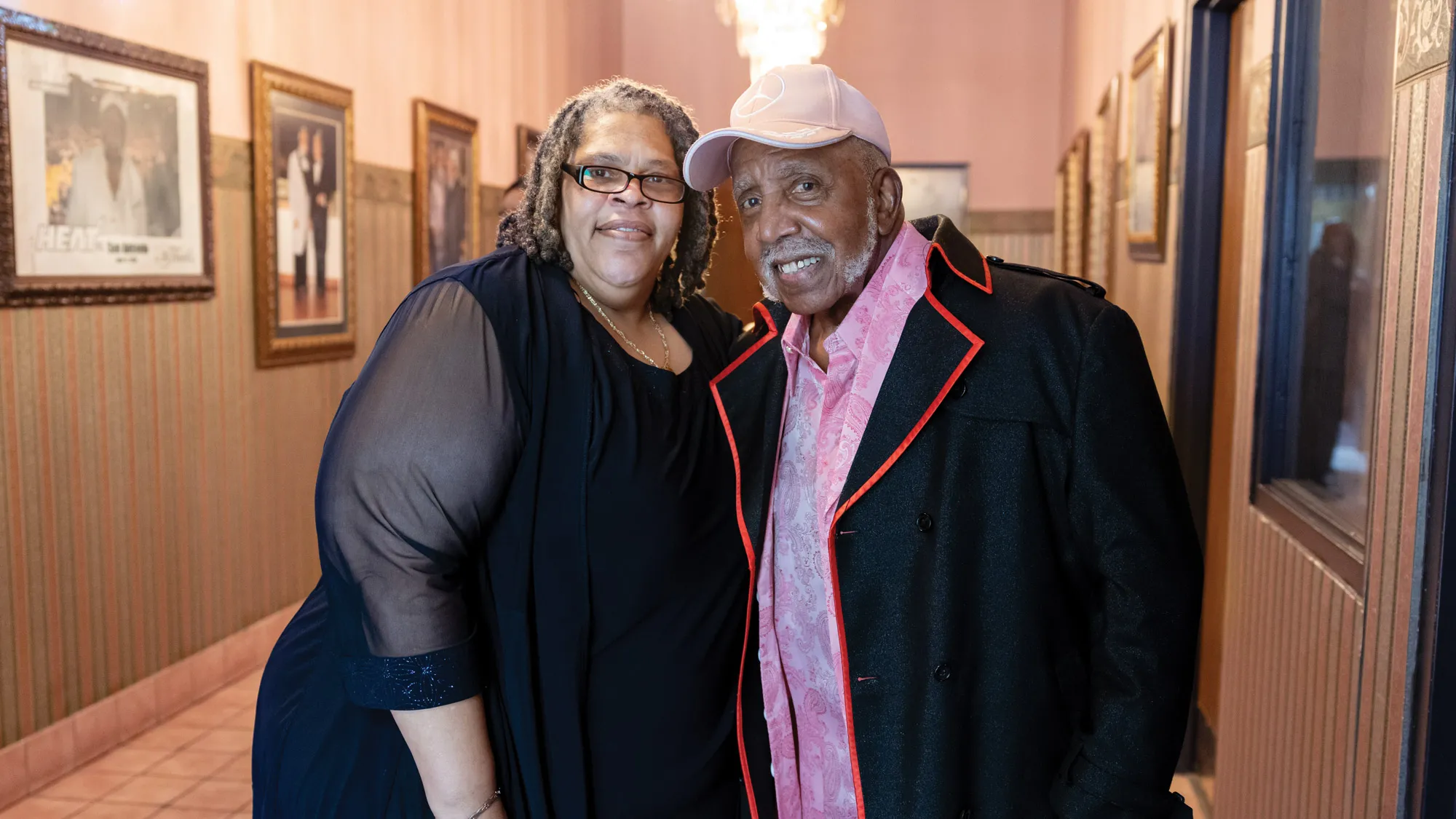 In a hallway lined with big framed photos, Palmer and her pastor pose for a photo. They’re both smiling, slightly leaning into each other, and they’re dressed for church. 