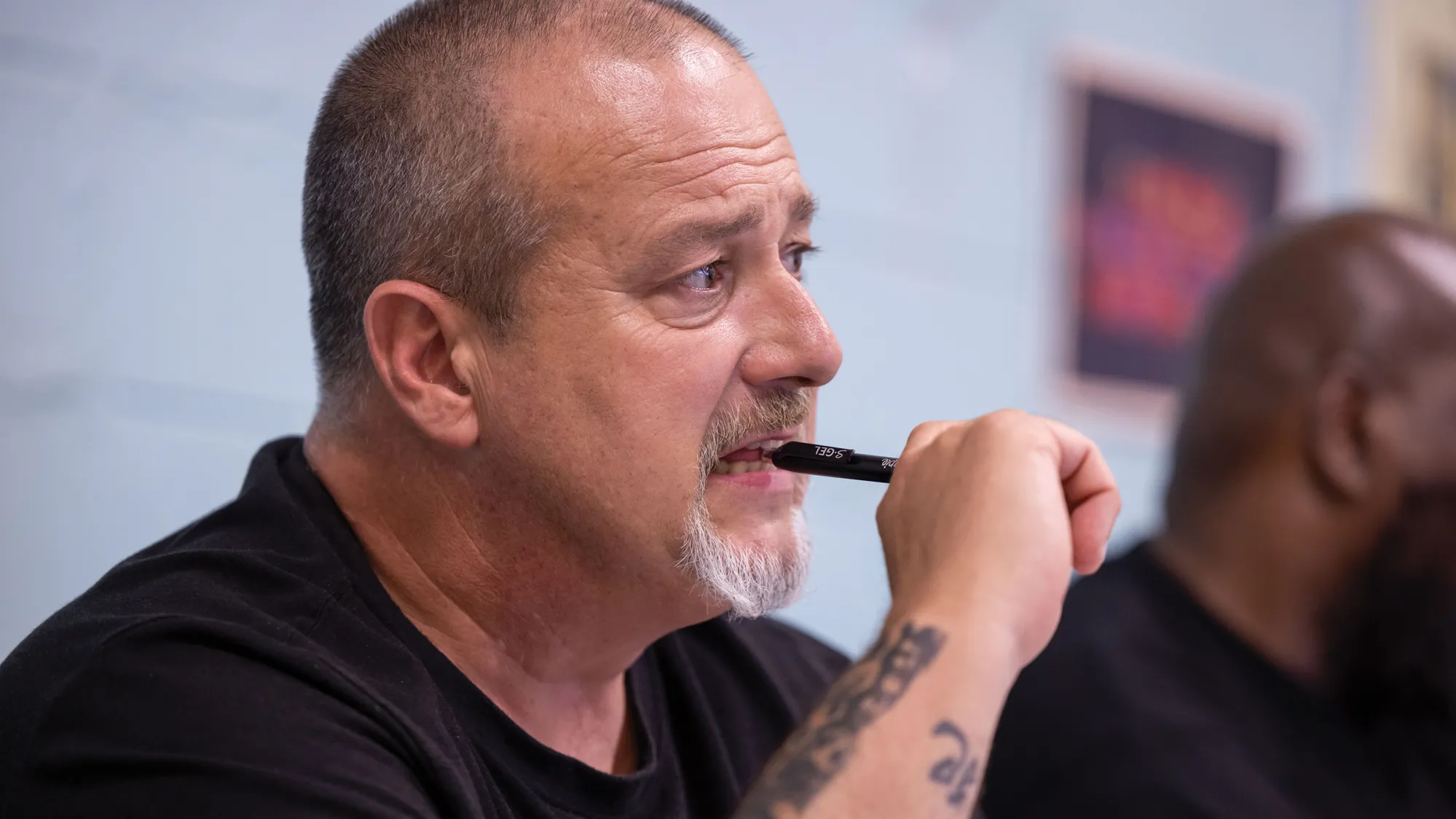 A man with tattoos on his arm bites on a pen as he listens in class.