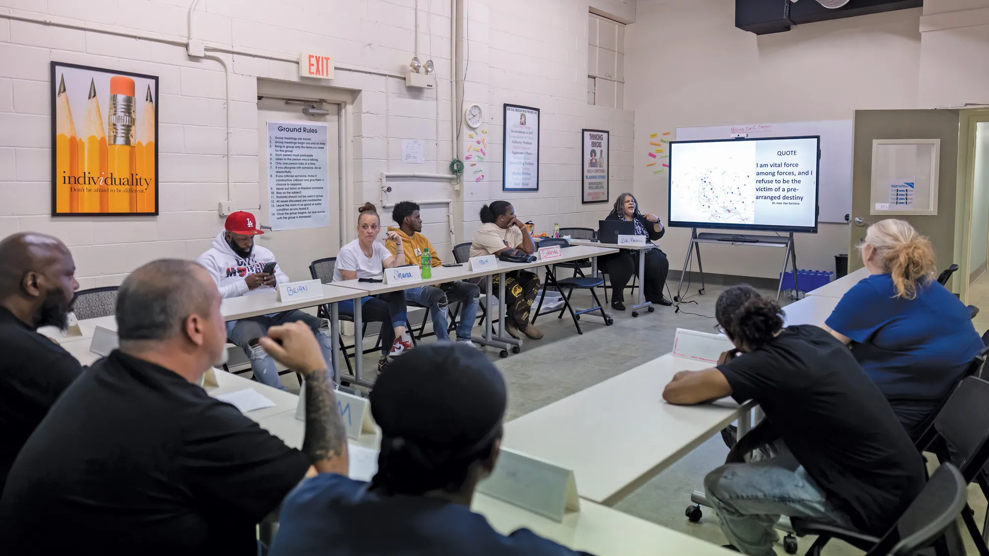 Students of various races, ages and genders sit at conference tables pulled into a U shape. Palmer sits at the front, talking as she presents on a screen.