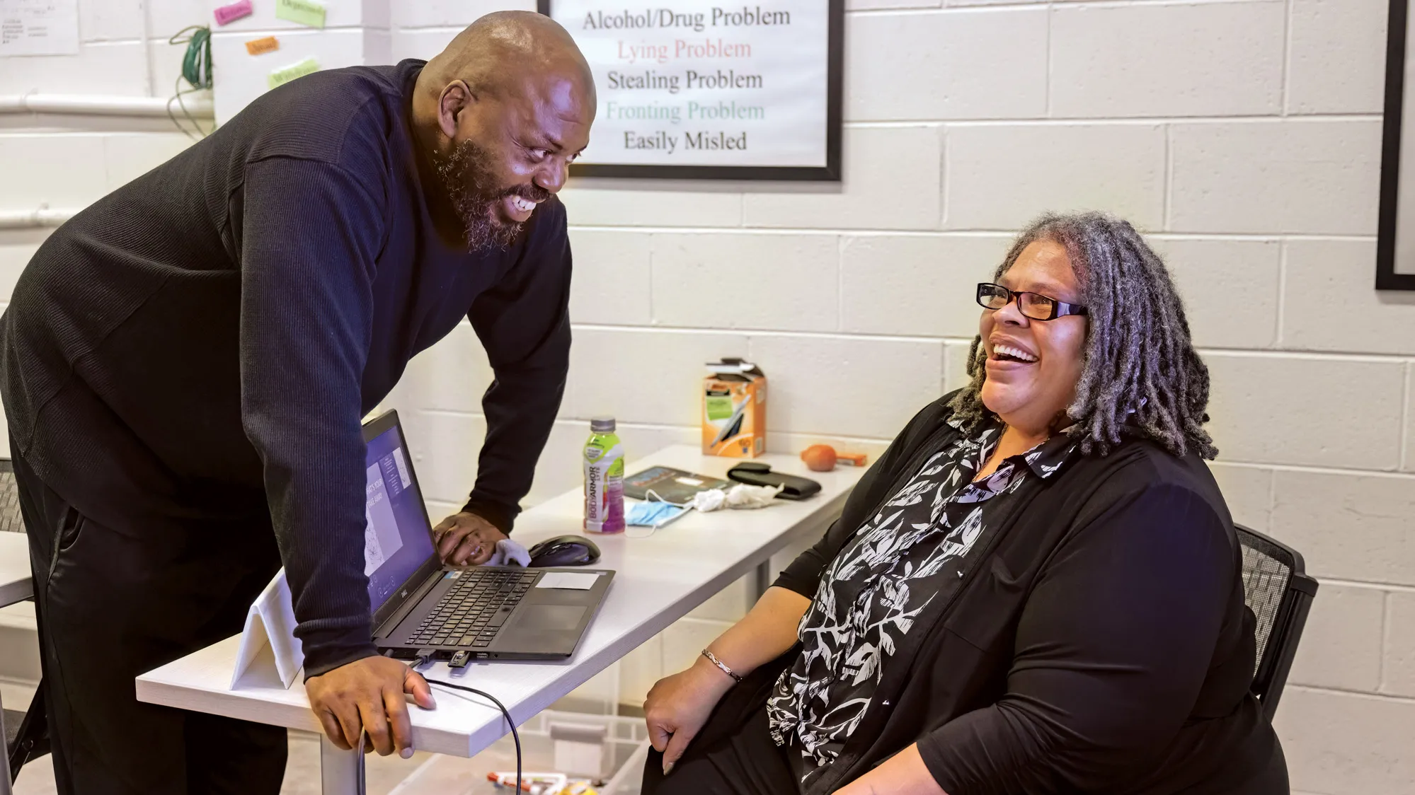 Patrice Palmer sits in a chair at a narrow table that holds her laptop and other things a teacher needs during class. She’s laughing as a man leans over the table and smiles as he watches her.