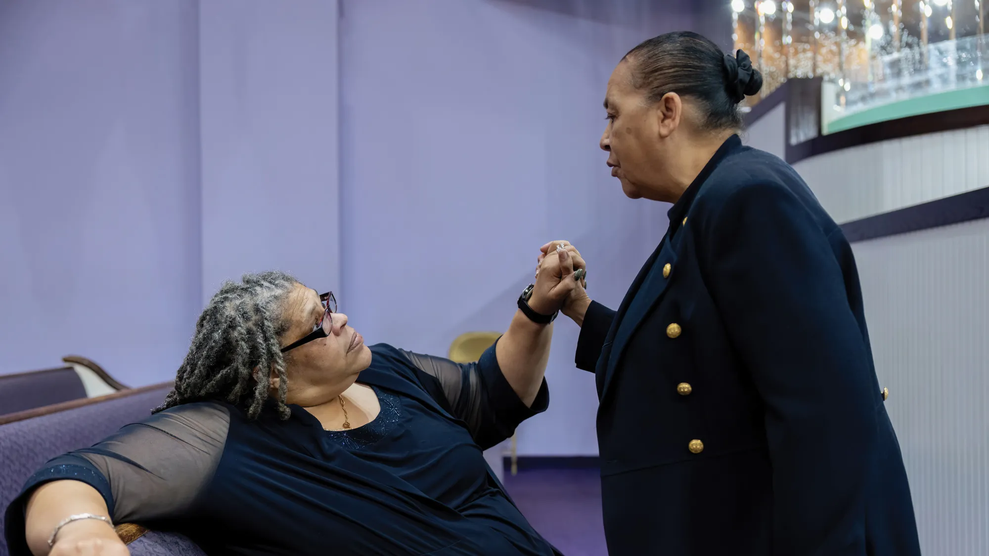 Sitting in an upholstered church pew, Palmer clasps hands with a woman standing in front of her. They’re making eye contact as they chat, and Palmer’s expression shows her actively listening. 