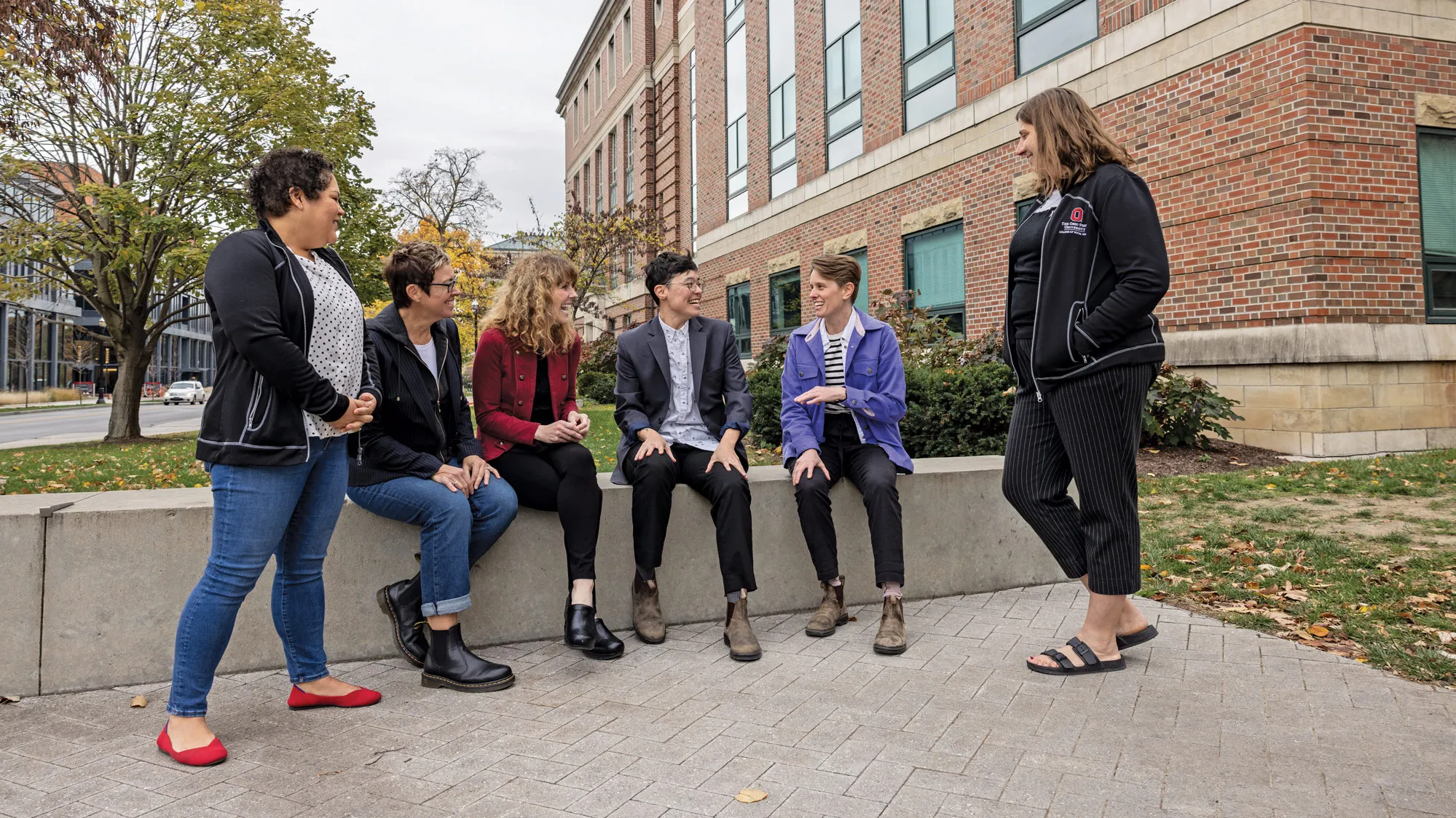 Outside of the brick Stillman Hall, a group gathers on a small paved plaza. Some sit on a low wall and some stand as they happily chat on what seems to be a chilly fall day, judging by their jackets and leaf colors on trees in the background. The people are a mix of ages and genders.