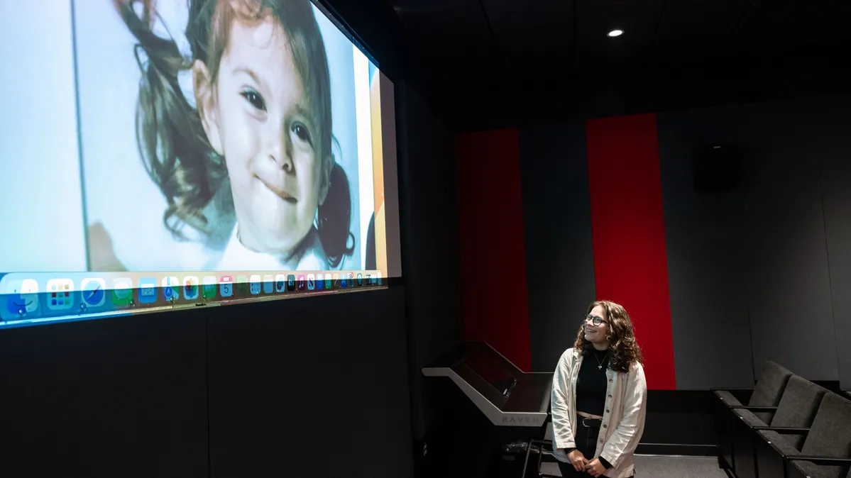A young woman clasps her hands in front of her as she stands at the front of a mini-movie theatre—the screening room in the new theatre building. She’s looking up at the movie screen, which is showing her documentary. The photo now on screen is her as a child of about 3 or 4.