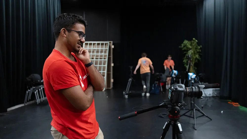 A young man stands in a thinking-liek pose as he considers the screen on the back of the camera he's using to film a practice scene. He's in a dark studio-walls and surrounding curtains are black and people work in the distant background.