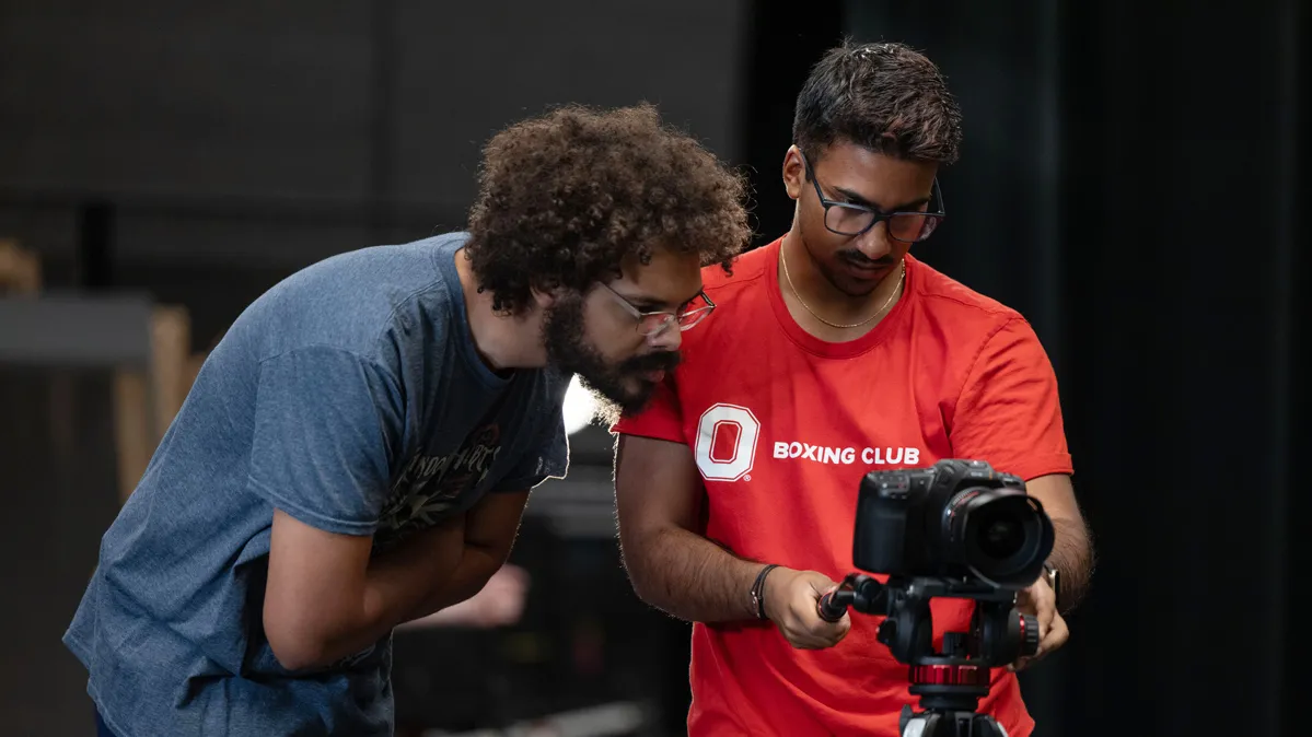 Two young men study how a scene is showing up on the screen on the back of their camera, which sits on a tripod that is almost chest-high.