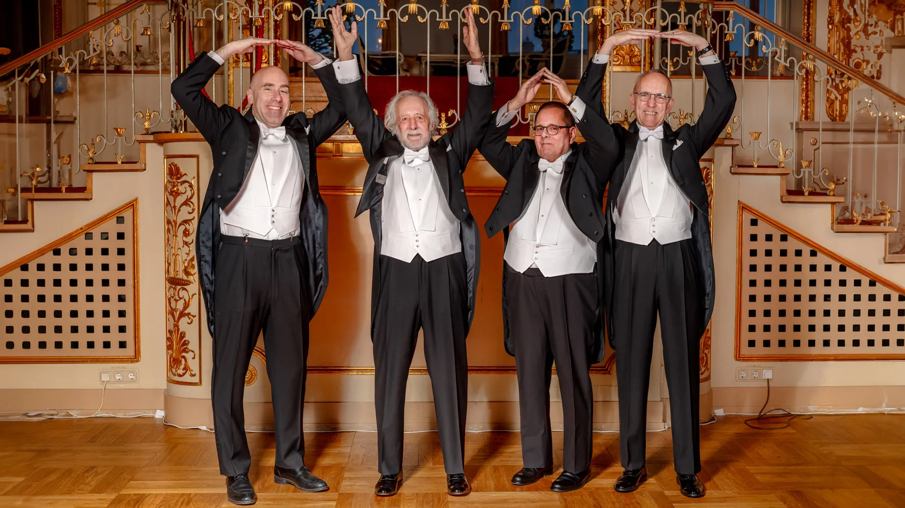 Standing in front of an ornate platform with stairs that extend from both sides, four white men dressed in tuxedos in matching color schemes each form a letter with their arms above their heads, spelling out OHIO. The second man, Nobel winner Pierre Agostini, has flowing white hair, a beard and a smile as he reaches high to make the H.
