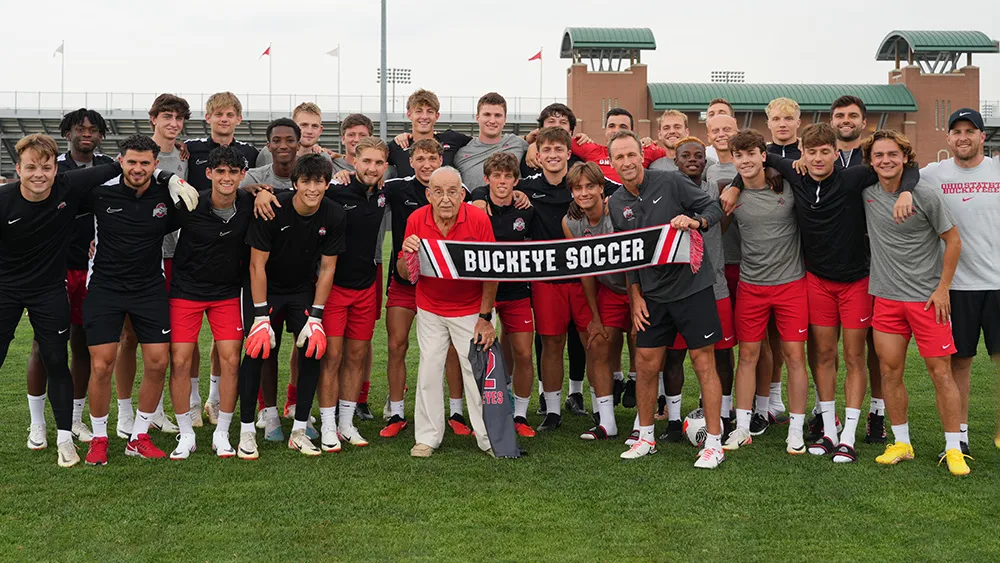 Paul Halpern, a 91-year-old man, poses with Ohio State's men's football team and coach on the soccer field at Schumaker Complex. They hold a banner that reads Buckeye soccer