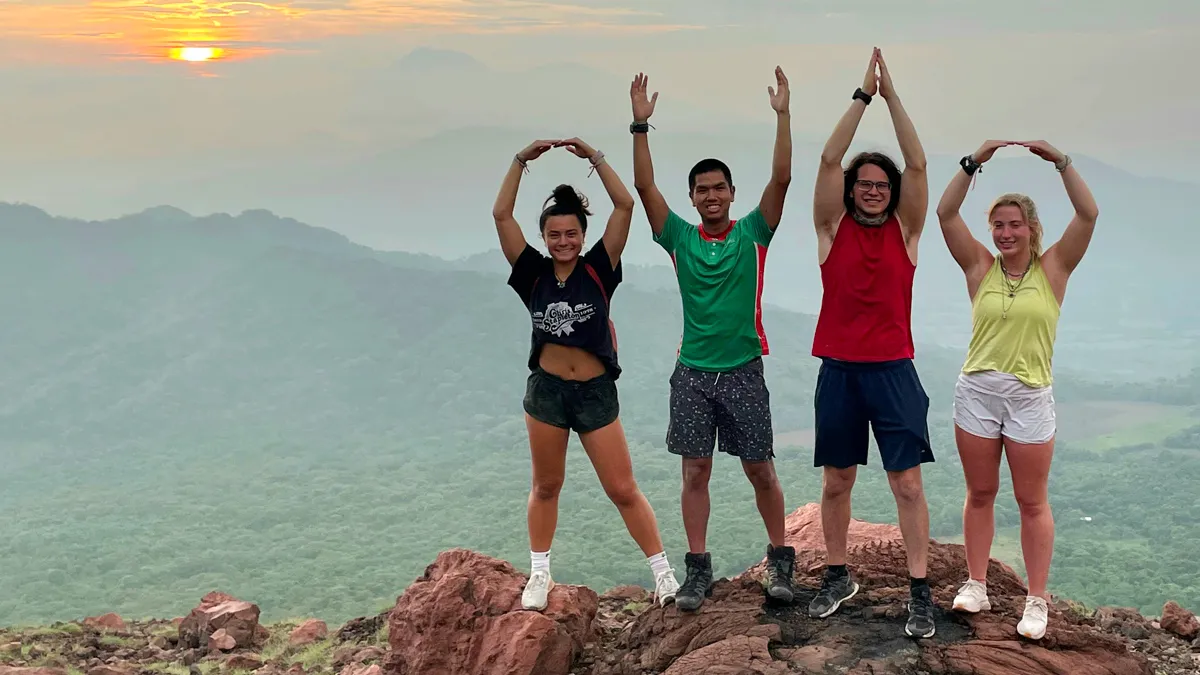 Standing on the edge of what seems to be a rocky cliff, four young adults spell out OHIO with their arms above their heads. They’re all wearing shorts, T-shirts and tennis shoes; those forming O’s are females, and the inside letters are formed by males. Behind them in the far distance is a valley coated in green trees and layers of mountains. The sun seems to be setting behind clouds.