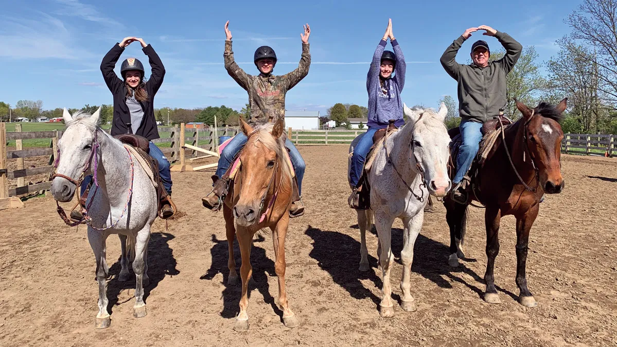 Four horses stand in a row facing the camera as their riders use their arms to spell out OHIO. They’re in a dirt paddock; beyond the fence is a grassy field and a barnlike building. The people are three girls and their dad.