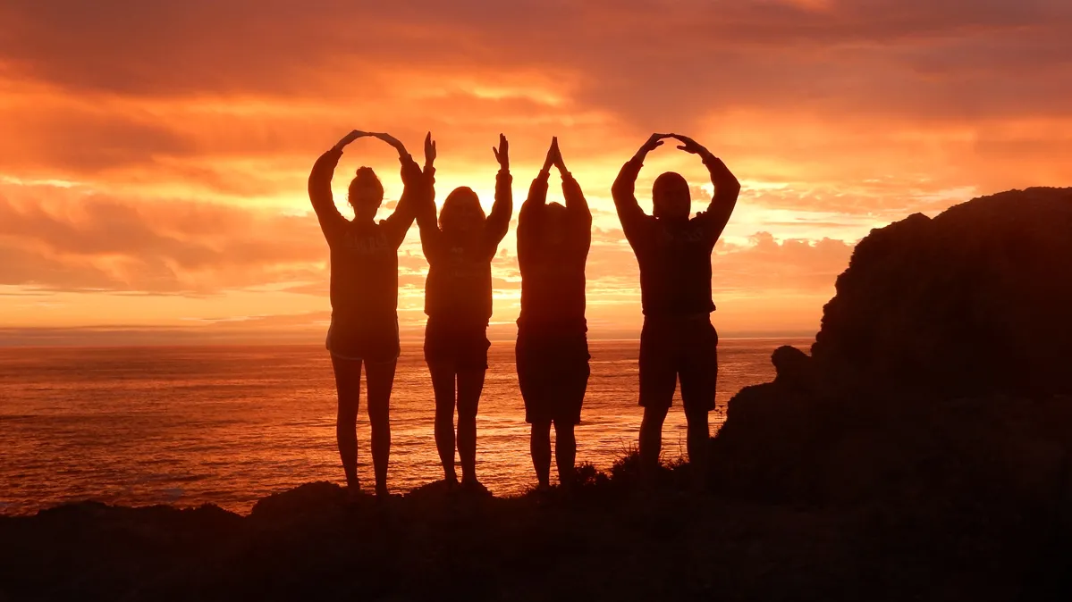 As the setting sun lights up the evening sky in shades of gold and orange, four people form an OHIO. Because of the backlighting, they appear solely as dark silhouettes.