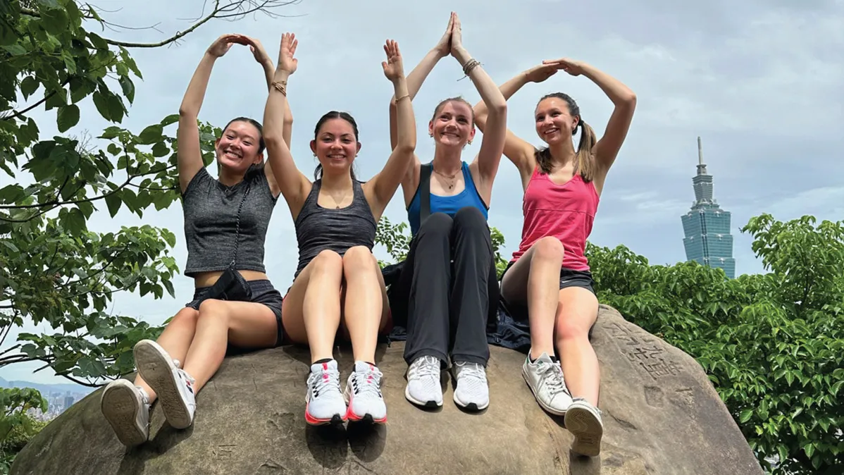 Sitting on a giant rock, four young women use their upper bodies to spell out OHIO as they smile happily. In the background a tall Taipei building made of glass rises above the treetops.