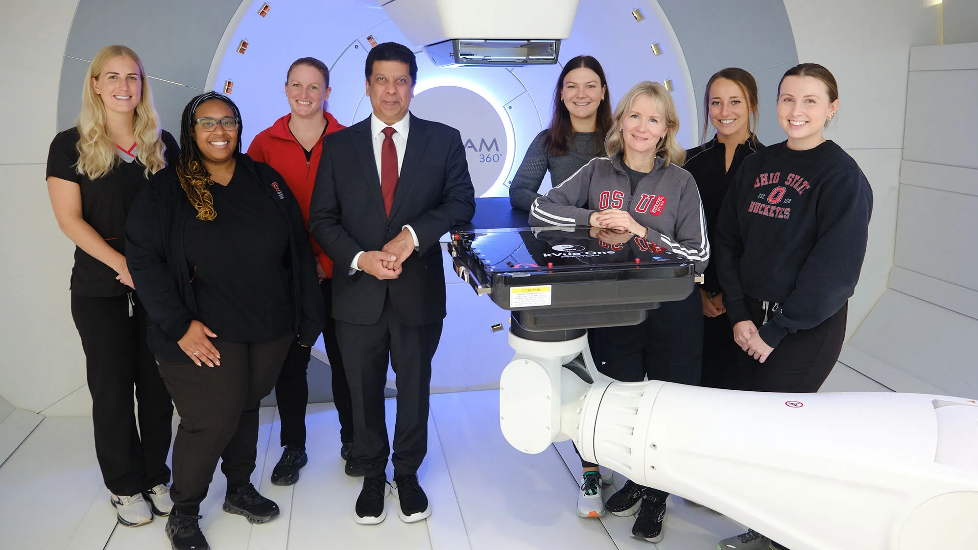 A team of people who work at Ohio State’s proton therapy center pose in the room where the treatment takes place. It is white-walled and appears spotless. They are eight smiling people—seven women—wearing scrubs or Ohio State sweatshirts. Dr. Arnab Chakravarti stands near the center in a suit.