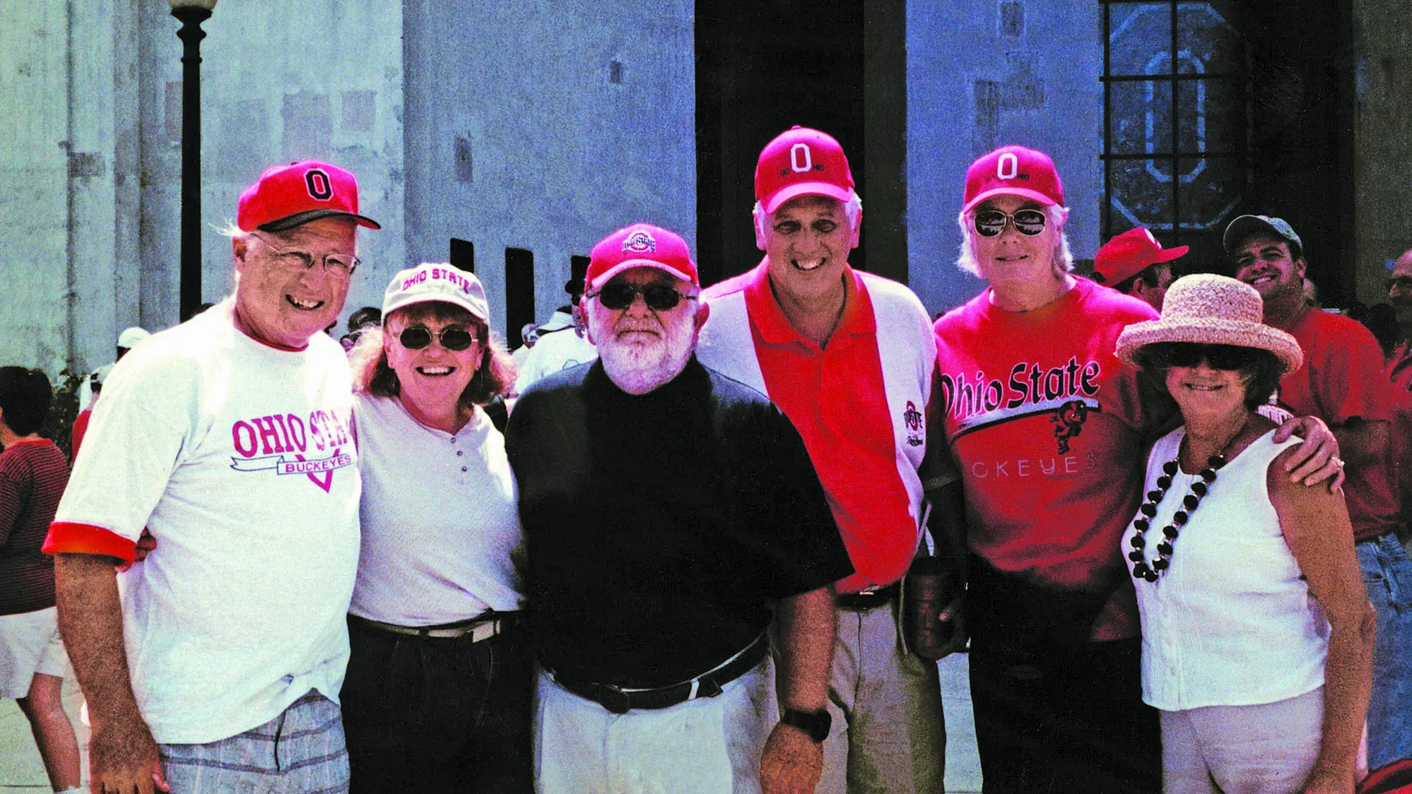 Three couples pose in front of the entrance to Ohio Stadium. They are all white, middle-aged people wearing Ohio State gear and baseball caps (though one lady has on a brimmed sunhat) who stand smiling with their arms around the shoulders of the people next to them.