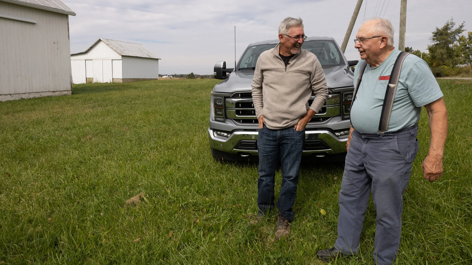 Two men laugh as they chat in a grassy area in front of a truck. Two painted barnlike buildings are off to their right. Both men are older white men. One, Doug Morgan, wears a quarter-zip sweater and jeans and has his hands tucked in his pockets as he listens. The other, Larry Layman, wears a T-shirt and work pants held up by suspenders and looks like he’s telling a good story. 
