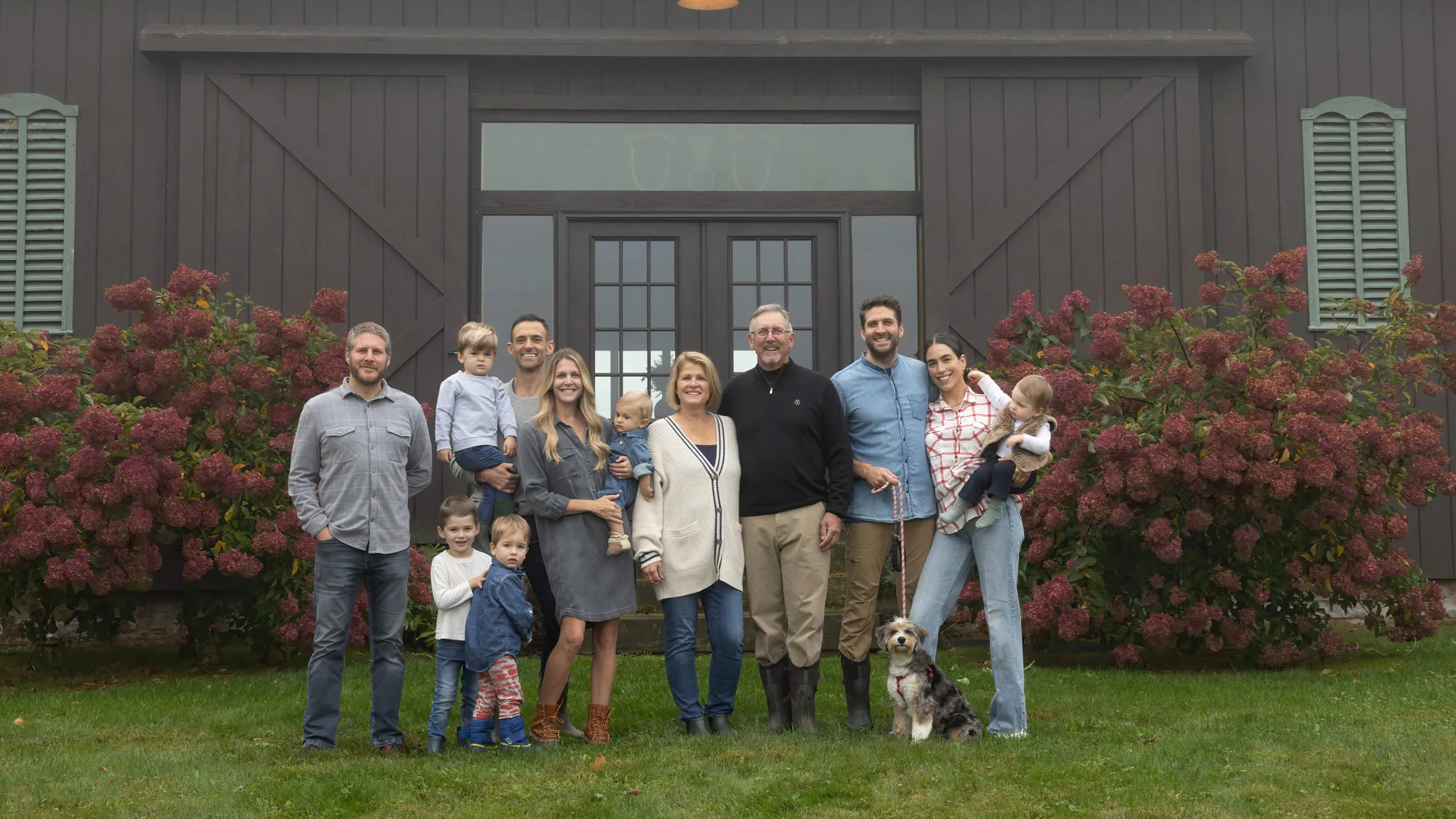 Doug and Beth Morgan stand in the middle as their family poses in front of a dark-stained barn. All the adults seem to be genuinely smiling. There are three other men, two other women, and four kids ranging from maybe 3 or 4 years old to 8 months. (They seem more puzzled  and distracted than posing!) And a shaggy, smaller sized dog on a leash. 